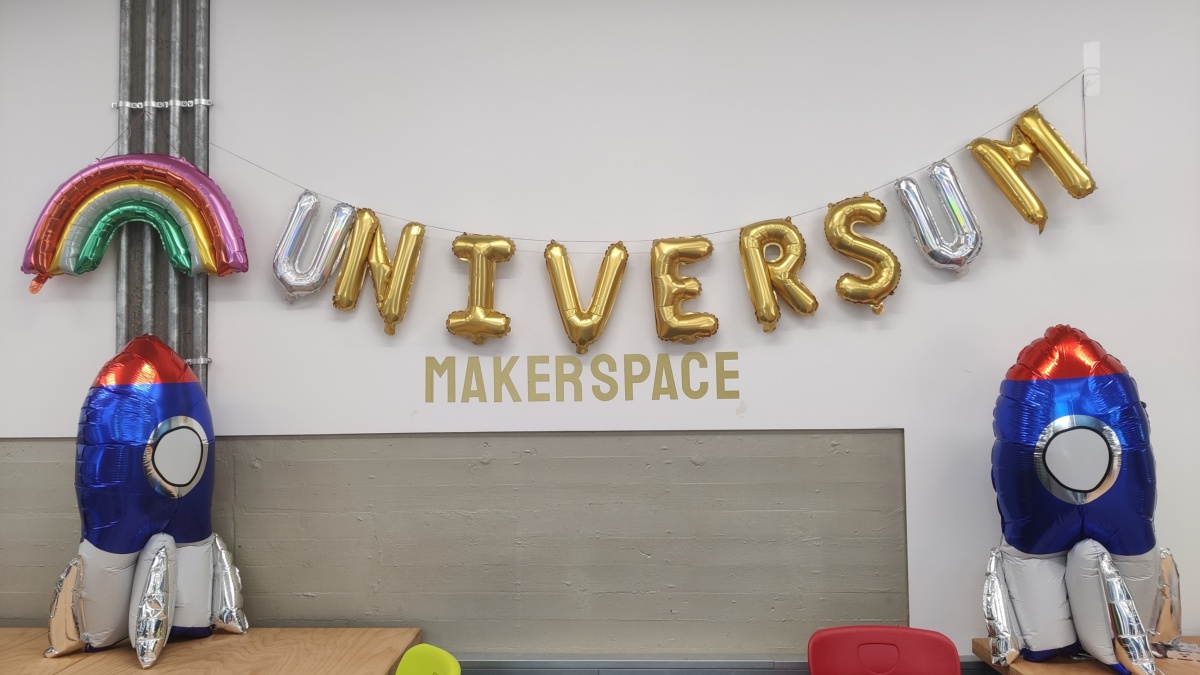 eine Ballongirlande schreibt das Wort "Universum" in goldenen Buchstaben, die "U"s sind aus Holo-Material. Links daneben ein Regenbogen-Ballon, rechts und links steht je ein großer blauer Raketen-Ballon.
