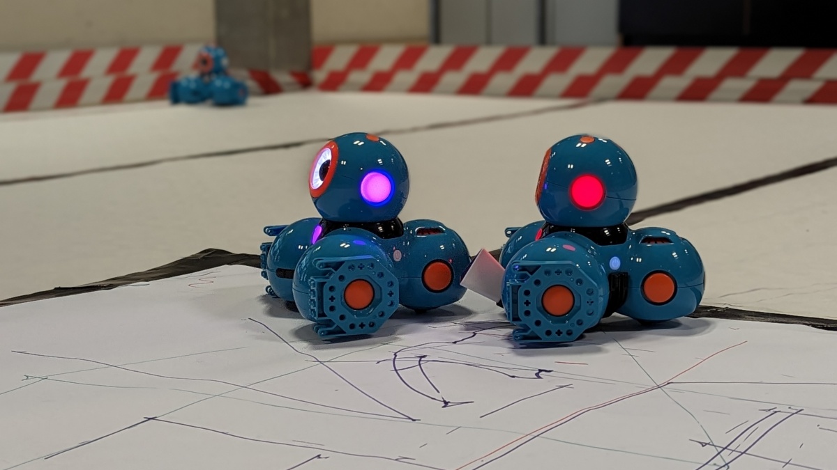 zwei kleine blaue Roboter aus Kugelelementen mit leuchtenden "Ohren" stehen auf einer Papierfläche, die sie gerade bemalt haben